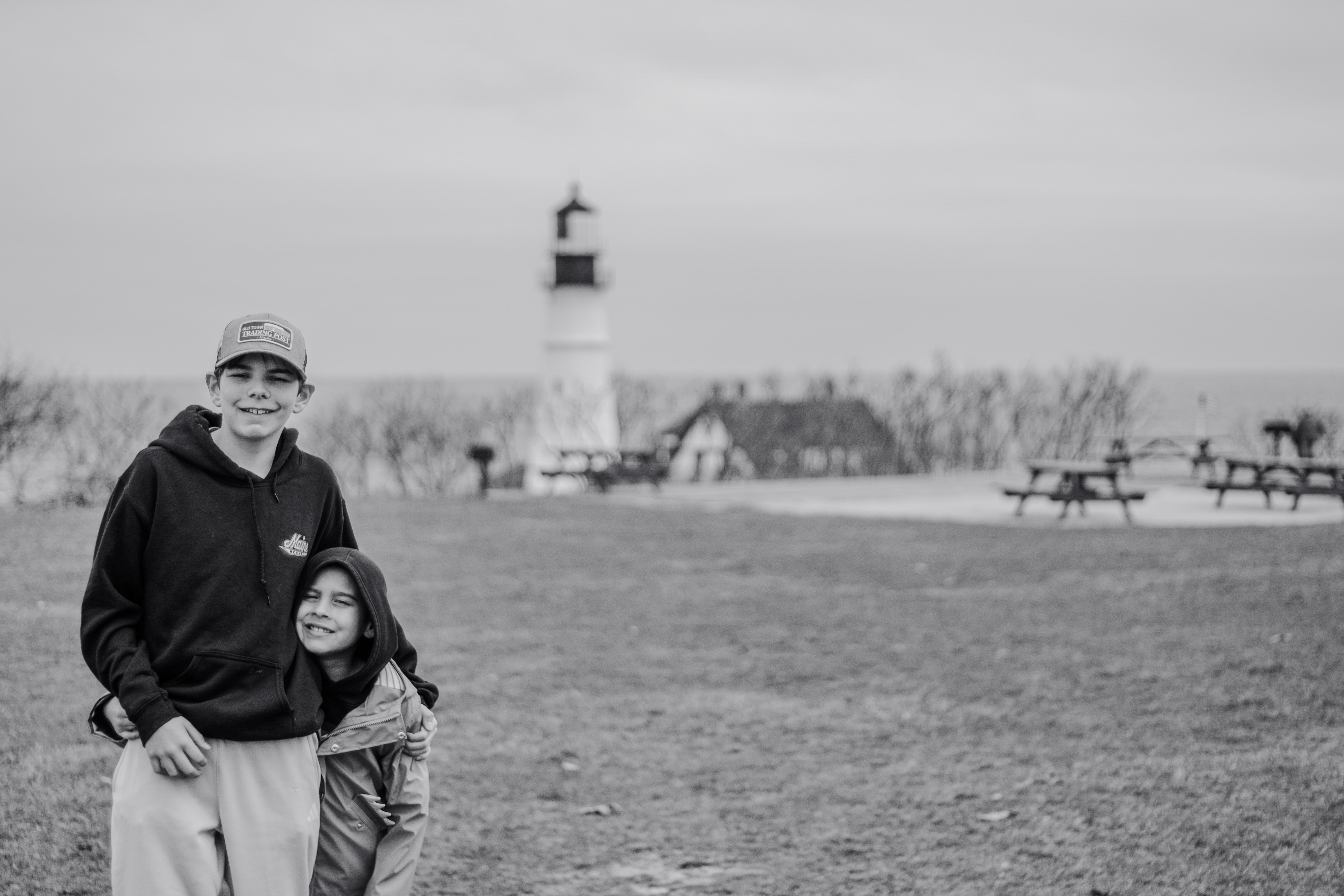 Black and white portrait of two siblings at a Maine lighthouse park, one hugging the other.